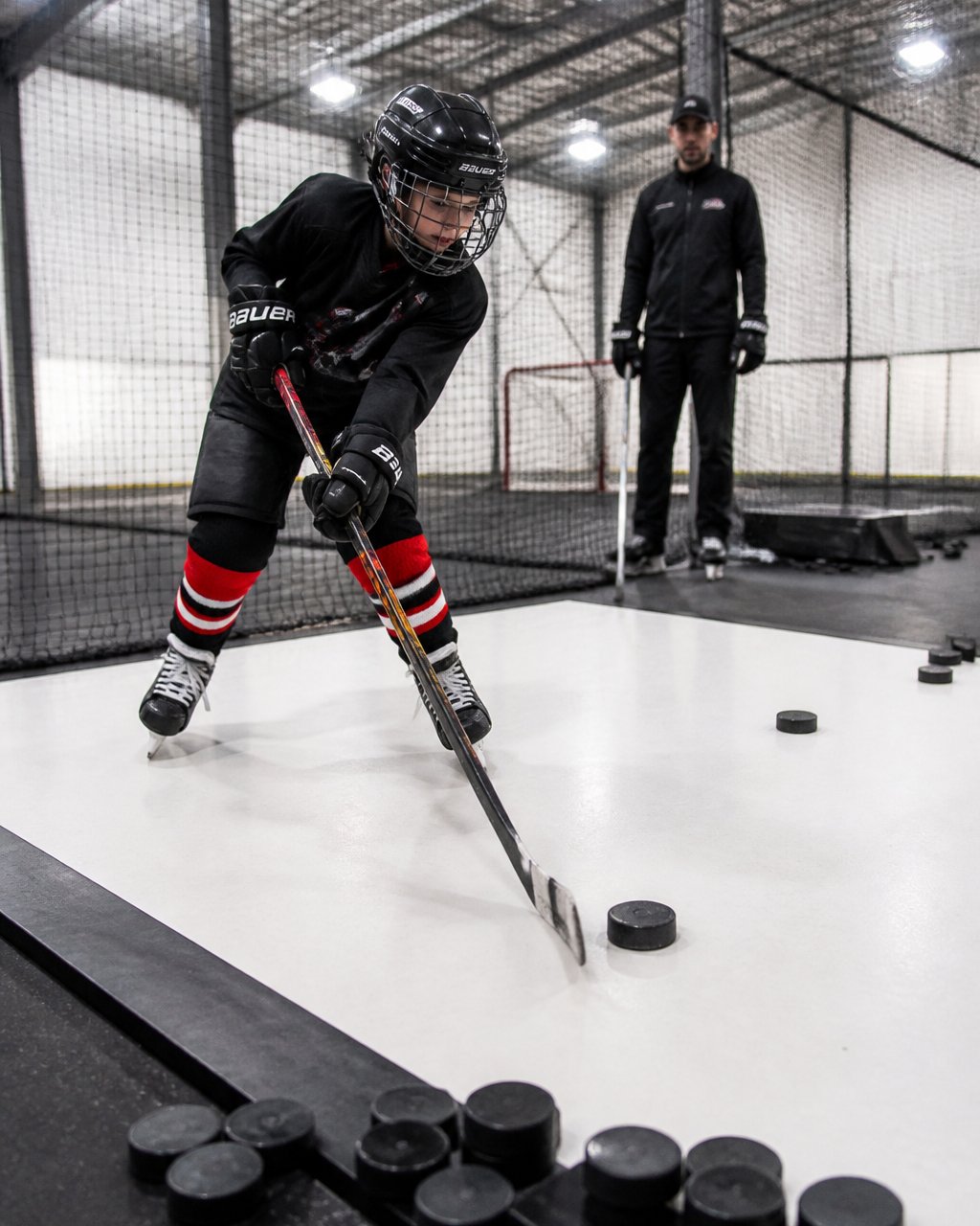 Young hockey player practicing wrist shots at indoor shooting lane facilities with a coach nearby.