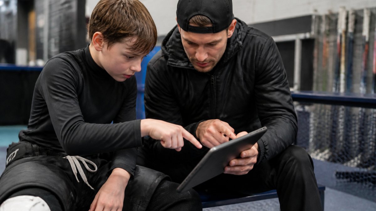 Young hockey player and coach reviewing game footage together on a tablet in a training facility, both pointing at the screen during analysis with focused concentration