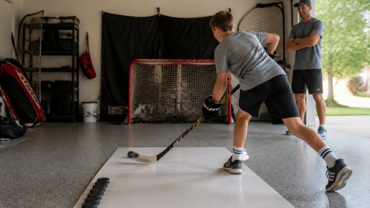 A young hockey player practicing wrist shots on a shooting pad in a garage training space, with pucks lined up and a net in the background, parent watching nearby