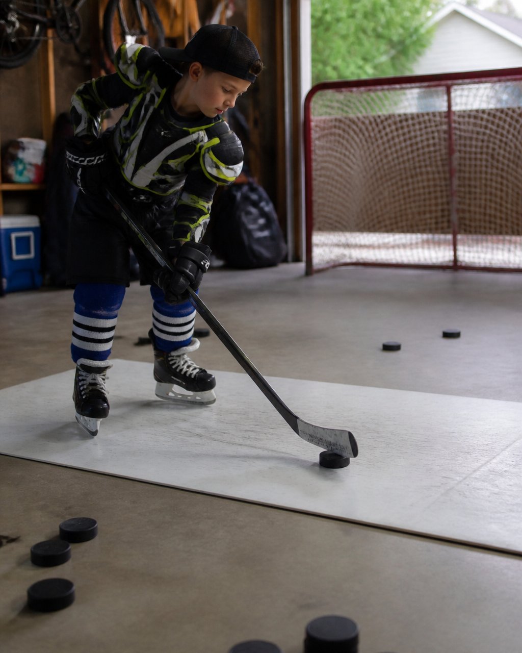A youth hockey player in full gear practicing wrist shots on a shooting pad in a garage, with a net and scattered pucks visible, focused expression, natural daylight from an open door